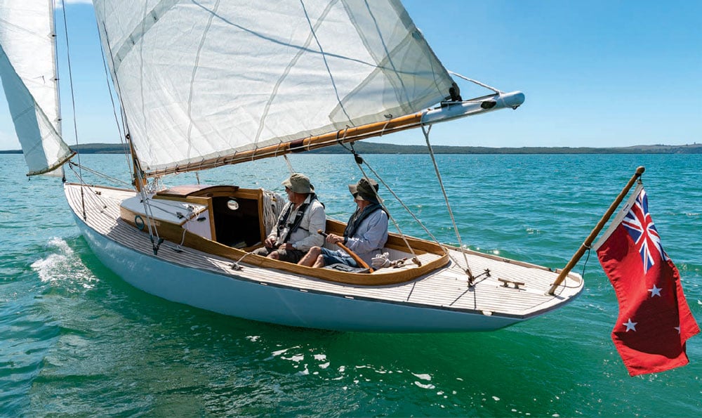 Two sailors aboard a small sailboat with a New Zealand Red Ensign flag.