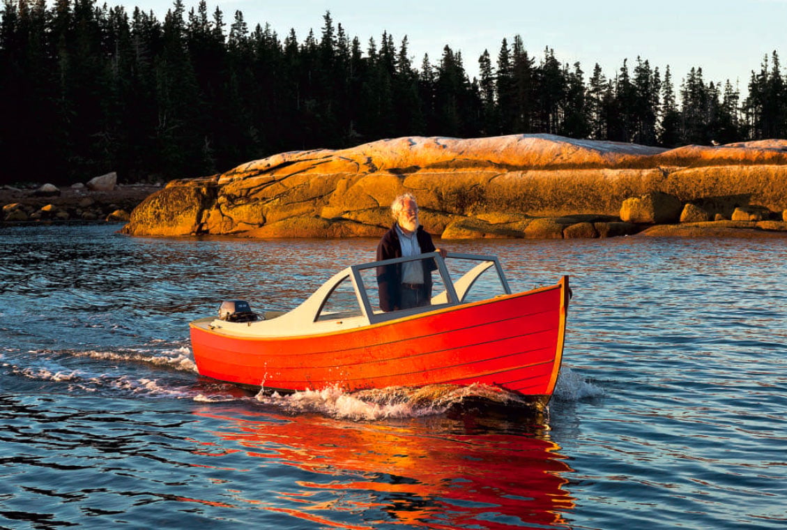 Man stands in a red powerboat on the water with pine trees and a rocky shore in the background.
