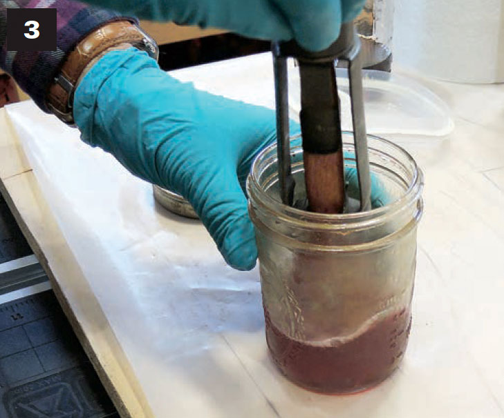 Gloved hands holding varnishing brushes in a glass jar of kerosene.