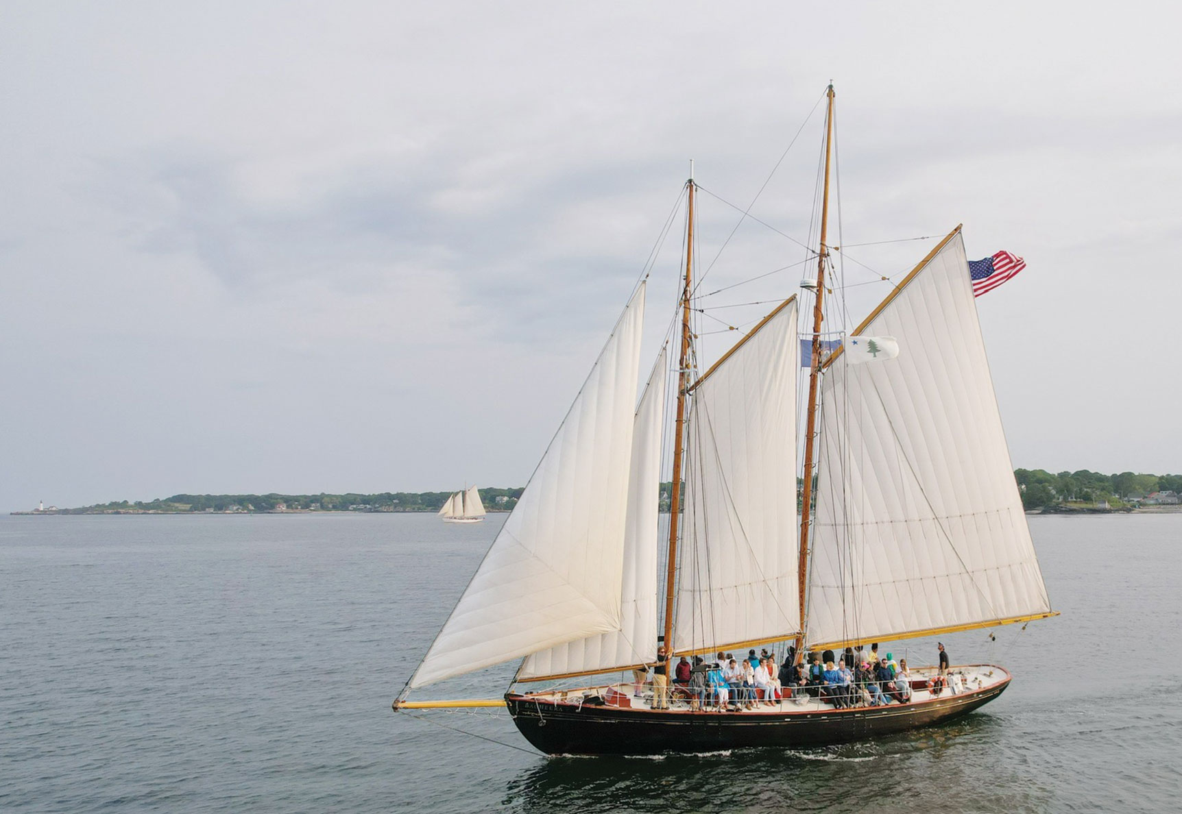 A group of people ride aboard the schooner BAGHEERA with a shoreline in the background.